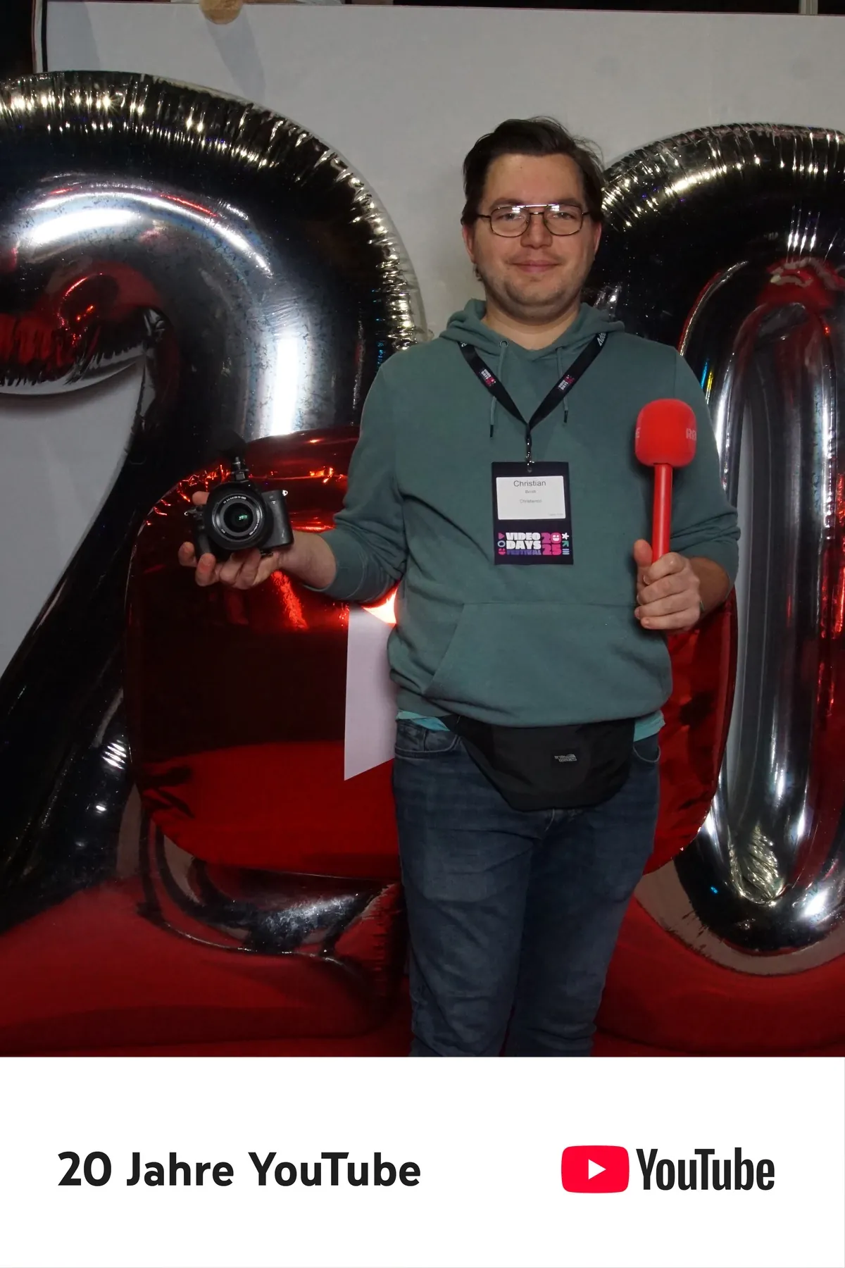 Christian mit Kamera und rotem Mikrofon vor der 20-Jahre-YouTube-Kulisse